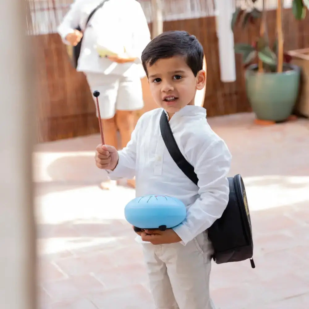 Un niño viste ropa blanca con un bolso negro al hombro y sostiene un contenedor azul.