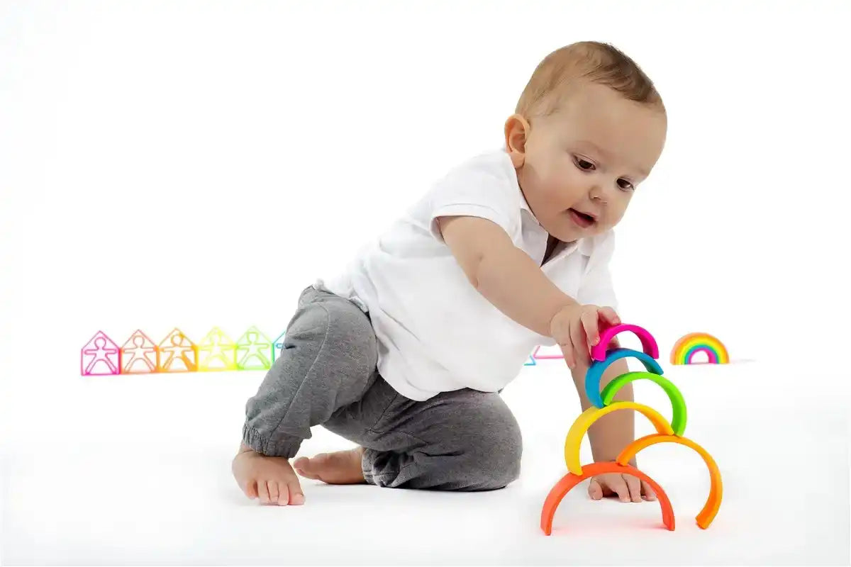 Bebé con camisa blanca y pantalones grises jugando con coloridos juguetes apilables en forma de arcoíris.